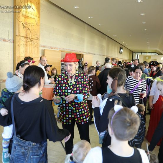 Eine Gruppe von Menschen in Kostümen bei einer Indoor-Veranstaltung. Eine Frau in einem gepunkteten Outfit mit rotem Hut steht in der Mitte, umgeben von anderen. Ein Schild oben zeigt das Datum 21.2.2023.