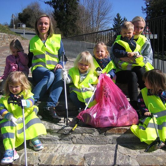 Eine Gruppe von Erwachsenen und Kindern, alle in gelben Westen, sitzen auf Steinstufen. Einige von ihnen haben Handschuhe und Stöcke. Sie scheinen einen Bereich aufzuräumen.