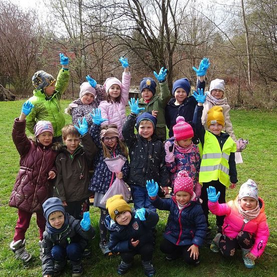 Eine Gruppe von Kindern in Winterkleidung posiert für ein Foto auf einer Rasenfläche. Sie tragen alle Handschuhe, einige haben Mützen. Die Kinder lächeln und heben ihre Hände.
