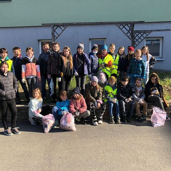 Eine Gruppe von Kindern in Winterkleidung posiert für ein Foto vor einem Gebäude, einige sitzen auf einer Bank und andere stehen und halten Plastiktüten.