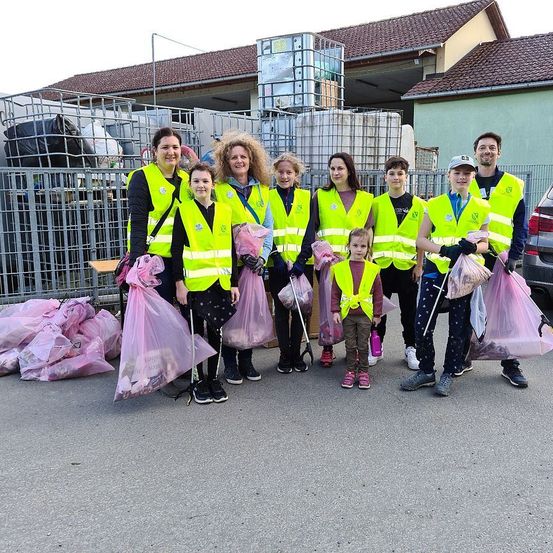 Eine Gruppe von Freiwilligen in gelben Sicherheitswesten und mit rosa Taschen posiert für ein Foto in der Nähe eines Metallzauns. Dahinter stehen ein Gebäude und ein Auto.