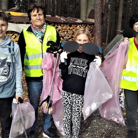Eine Gruppe von Menschen, darunter zwei Frauen, ein Junge und ein Mädchen, stehen in einem Wald. Sie halten Plastiktüten, Stöcke und Masken in den Händen. Hinter ihnen befinden sich Holzstapel.