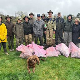 Eine Gruppe von Menschen in Outdoor-Ausrüstung, einschließlich Hüten und Stiefeln, steht auf einem Grasfeld mit Säcken voller gesammelter Gegenstände. Ein brauner Hund liegt auf dem Gras, und einige Leute halten grüne Flaschen. Bäume und ein bewölkter Himmel sind im Hintergrund zu sehen.