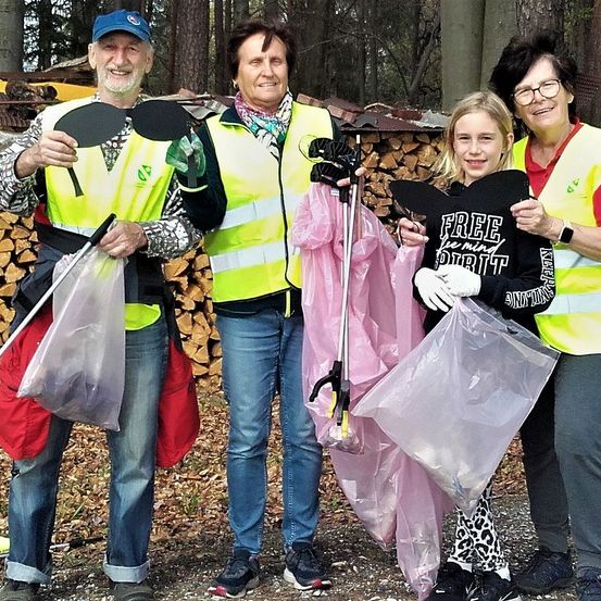 Vier Menschen, zwei Männer und zwei Frauen, stehen im Freien und halten Plastiktüten. Sie tragen Warnwesten und Handschuhe. Hinter ihnen ist ein Haufen Holz zu sehen, und der Hintergrund ist mit Bäumen gefüllt.