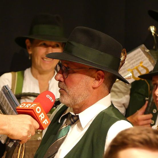 Ein Mann wird von einem Reporter mit einem Mikrofon interviewt. Der Mann trägt einen Hut und eine Brille. Hinter ihm steht eine Frau, die ebenfalls einen Hut trägt und lächelt. Im Hintergrund sind weitere Personen in Hüten zu sehen.
