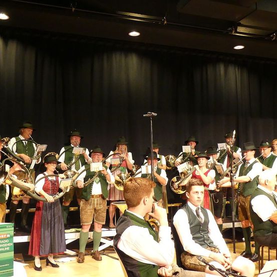 Eine Gruppe von Musikern in traditioneller bayerischer Kleidung spielt auf der Bühne mit verschiedenen Blechblasinstrumenten, während einige Zuschauer von vorne zusehen.