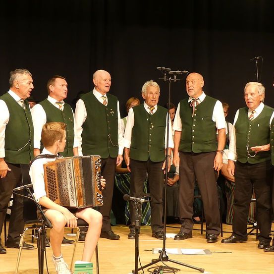 Eine Gruppe älterer Männer in grünen Westen singt auf der Bühne, während ein junger Junge ein Akkordeon spielt, der vor ihnen sitzt.