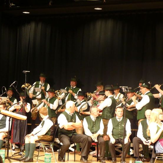 Eine Gruppe von Männern und Frauen in traditioneller grüner und weißer Kleidung spielt auf der Bühne mit Blech- und Holzblasinstrumenten, während einige sitzen und zusehen.
