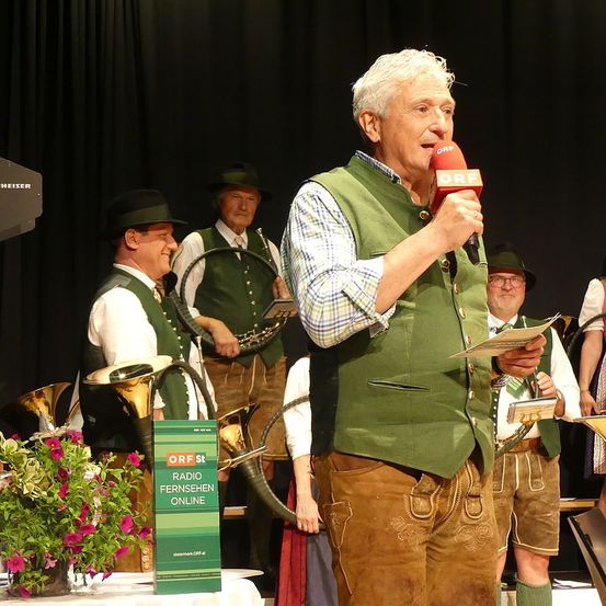 Ein älterer Mann steht auf der Bühne, hält ein Mikrofon in der Hand und spricht, während eine Gruppe von Menschen in traditioneller deutscher Kleidung hinter ihm steht. Sie halten Musikinstrumente in den Händen. Ein Schild mit dem Text 'ORF St Radio Fernsehen Online' steht vor der Bühne.