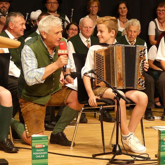 Ein Mann mit einem Mikrofon interviewt einen jungen Jungen, der auf der Bühne ein Akkordeon spielt, während eine Menschenmenge in traditioneller Kleidung im Hintergrund steht.