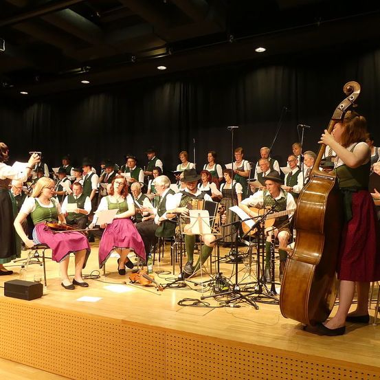 Eine Gruppe von Musikern, die auf der Bühne mit verschiedenen Instrumenten auftreten. Einige sitzen, andere stehen, darunter ein Cellist. Das Publikum ist nicht sichtbar.