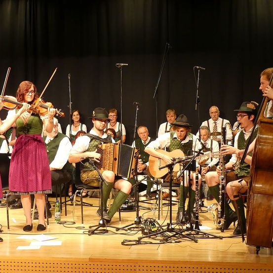 Eine Band tritt auf der Bühne auf, wobei eine Frau die Geige spielt und ein Mann das Akkordeon spielt. Andere Bandmitglieder spielen Gitarre, Trompete und Cello. Sie sind in traditioneller Kleidung gekleidet.