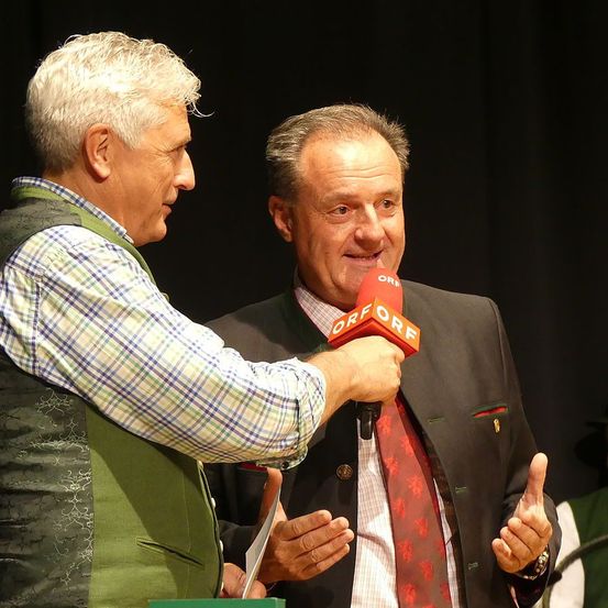 Ein älterer Mann wird von einem anderen Mann interviewt, der ein Mikrofon in der Hand hält. Der Interviewer trägt ein kariertes Hemd, während der Befragte einen Anzug und eine Krawatte trägt. Beide Männer lächeln und geben Daumen hoch. Der Befragte hält ein Stück Papier in der Hand. Das Mikrofon hat den Text 'ORF' darauf. Hinter ihnen ist ein schwarzer Vorhang.