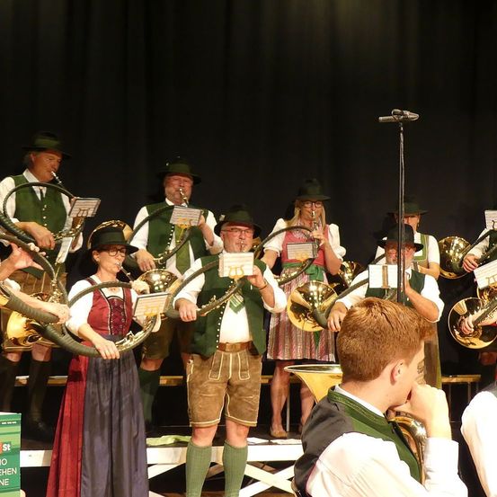 Eine Blaskapelle spielt auf der Bühne, wobei die Musiker verschiedene Blechblasinstrumente spielen. Ein Mann sitzt vorne und hört aufmerksam zu. Die Bandmitglieder tragen traditionelle bayerische Kleidung, einschließlich grüner Westen und Hüte.