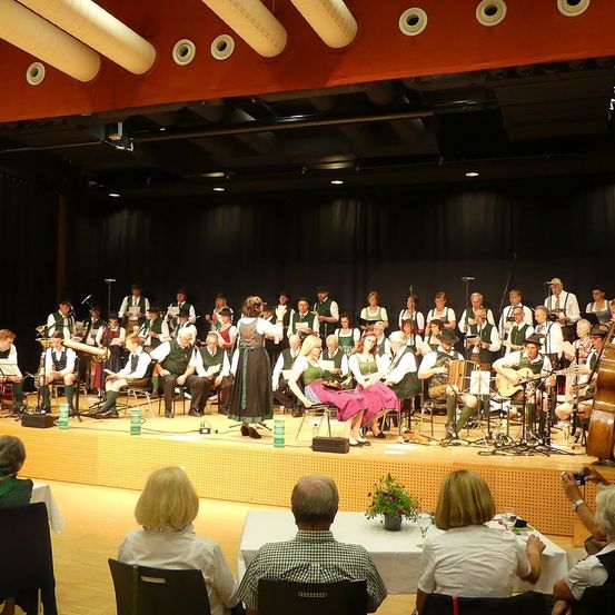 Eine große Gruppe von Menschen in traditioneller Kleidung führt auf der Bühne auf, mit Instrumenten wie Gitarren, Akkordeons und einer Geige. Das Publikum sitzt vor der Bühne an Tischen und beobachtet die Aufführung.