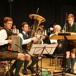 Eine Gruppe von Musikern spielt auf der Bühne. Einer spielt Tuba, ein anderer Akkordeon und ein Junge steht neben einem Mikrofon.