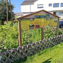Ein hölzernes Informationsschild mit gelben Plakaten zum Bienenschutz steht in einem Garten mit verschiedenen Pflanzen und Blumen. Hinter dem Schild befindet sich ein weißes Gebäude mit mehreren Fenstern.