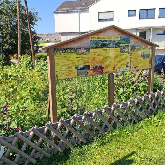 Ein hölzernes Informationsschild mit gelben Plakaten zum Bienenschutz steht in einem Garten mit verschiedenen Pflanzen und Blumen. Hinter dem Schild befindet sich ein weißes Gebäude mit mehreren Fenstern.