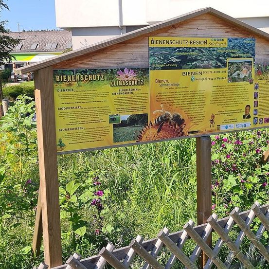 Ein Lehrtafel über Bienenschutz steht in einem Garten, umgeben von hohem Gras und Blumen. Das Schild ist gelb mit schwarzem Text und Illustrationen. Es ist an einer Holzkonstruktion befestigt.