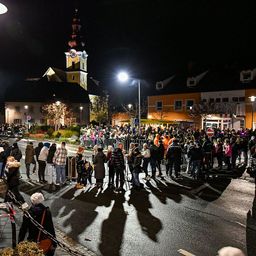 In der Nacht versammelt sich eine große Menschenmenge auf einer Straße mit Gebäuden und Straßenlaternen. Im Hintergrund ist eine Kirche mit einem Glockenturm zu sehen.