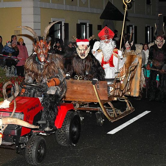 Eine Gruppe von Menschen in Kostümen, darunter einer als Weihnachtsmann verkleidet, fährt auf einem roten Traktor, der einen Schlitten zieht. Sie sind von Zuschauern im Hintergrund umgeben.