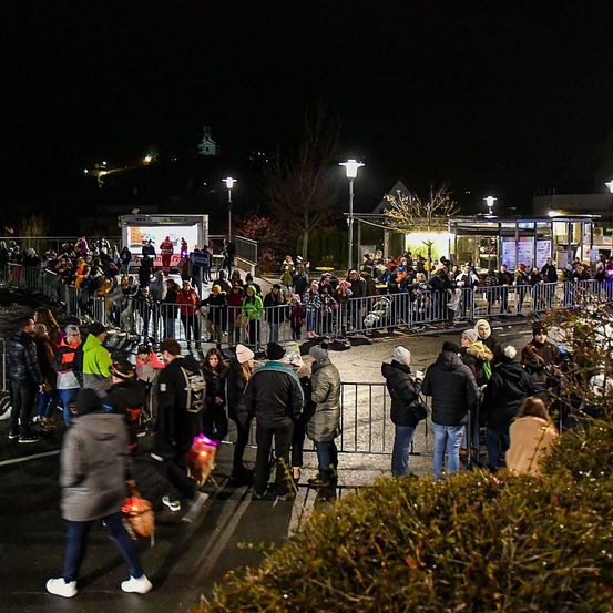 Eine große Menschenmenge versammelt sich bei Nacht im Freien, in Winterkleidung und mit Gegenständen. Sie stehen hinter Absperrungen in einer Reihe, einige stehen in der Nähe von Straßenlaternen. Das Gebiet ist von den Lichtern erhellt.