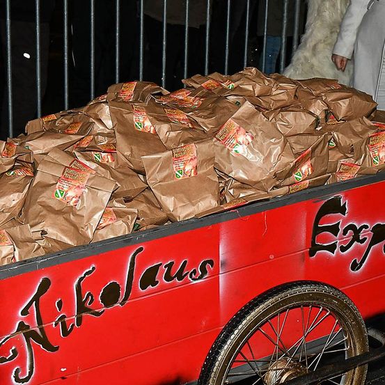 Ein roter Wagen voller brauner Papiertüten, beschriftet mit 'Nikolau' und 'Exp', steht mit Menschen hinter einem Metallzaun.