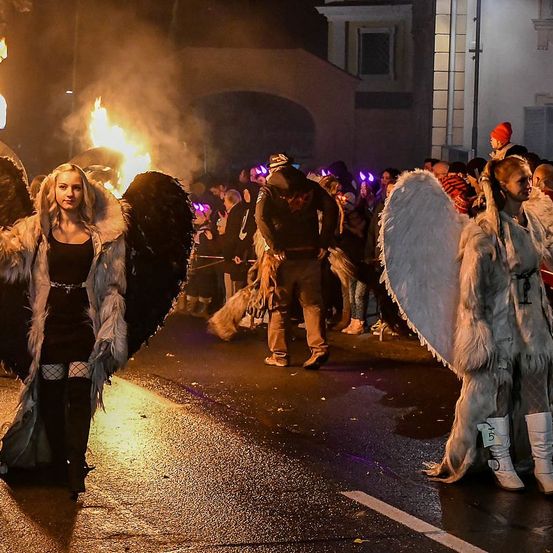 Ein nächtliches Festival mit einer Menschenmenge in Kostümen, zwei Frauen mit Engelsflügeln und einem Feuer im Hintergrund.