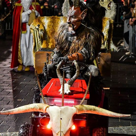 Eine Person, verkleidet als Teufel, sitzt in einem roten Traktor mit Kuhhörnern, einem Schädel und roten Lichtern hinten. Hinter ihr steht eine Person in einem Weihnachtsmann-Kostüm, während eine Menge zuschaut.
