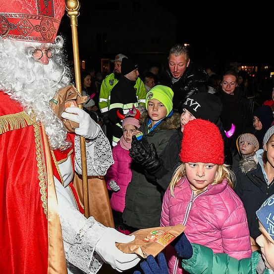 Der Weihnachtsmann in einem roten Gewand verteilt Geschenke an Kinder in einer Menschenmenge. Hinter ihm steht ein Mann in einer gelben Weste.