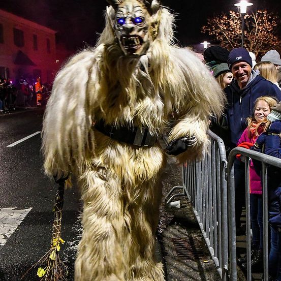 Eine Person in einem Monsterkostüm mit blauen Augen und einem breiten Mund geht bei Nacht in einer Parade und hält eine Besen. Dahinter steht eine Menschenmenge hinter einer Metallbarriere und beobachtet.