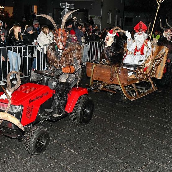 Eine Person in einem Ziegenkostüm fährt einen Traktor in einer Parade, gefolgt von einem Schlitten mit einer Person in einem Weihnachtsmannkostüm. Viele Menschen beobachten hinter einem Zaun.