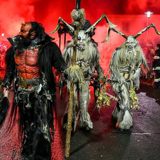 Drei Menschen in Kostümen stehen auf einer Straße, möglicherweise während eines Festes. Sie tragen gehörnte und haarige Outfits. Die Person links trägt ein rotes Muskelshirt. Ein Feuerwerksdisplay beleuchtet den Hintergrund.