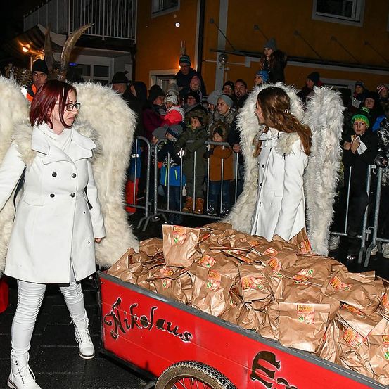 Zwei Frauen, die als Engel gekleidet sind, schieben einen roten Wagen voller Papiertüten durch eine Menge bei Nacht.