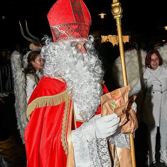 Ein Mann, der als Weihnachtsmann verkleidet ist, hält eine Tüte mit Süßigkeiten in der Hand, trägt einen roten Bischofshut und einen langen Stab. Er ist von Menschen in Kostümen umgeben, darunter eine Person mit Engelsflügeln. Der Hintergrund ist dunkel, mit Lichtern und Gebäuden in der Ferne sichtbar.