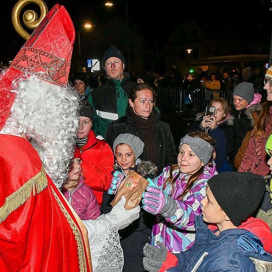 Eine Menschenmenge, darunter Kinder, versammelt sich um einen als Weihnachtsmann verkleideten Mann, der Geschenke verteilt. Die Szene spielt sich draußen bei Nacht ab.