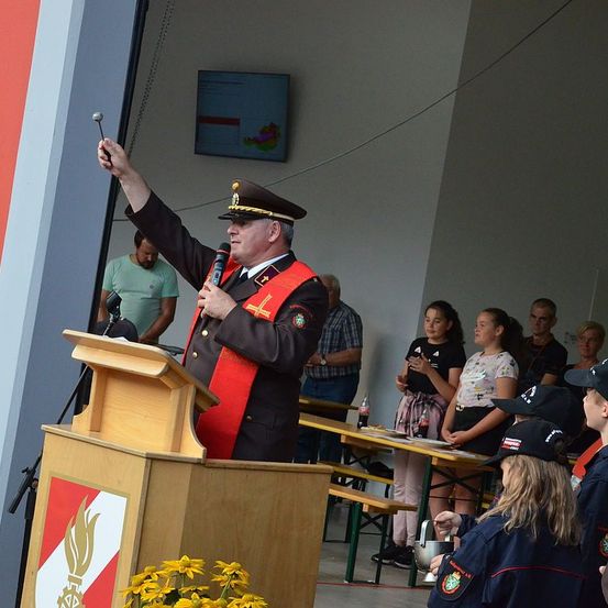 Ein Mann in Uniform steht an einem Podium und hält eine Rede mit einem Mikrofon. Hinter ihm sitzen und stehen mehrere Menschen, darunter eine Gruppe von Kindern. An der Wand ist ein Monitor angebracht.