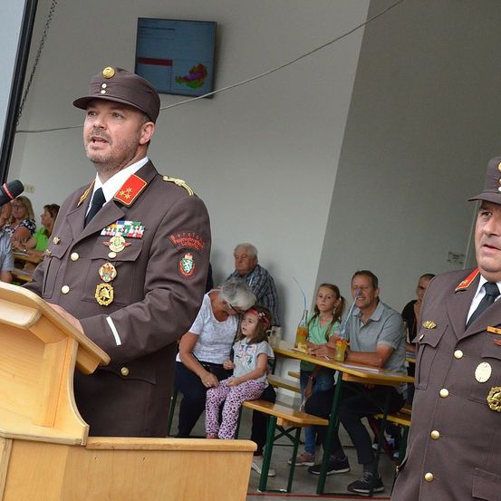 Zwei Männer in Militäruniformen stehen hinter einem Podium. Einer spricht in ein Mikrofon. Hinter ihnen sitzen Menschen an Tischen, darunter ein Mädchen mit Zöpfen. Eine andere Frau beobachtet den Redner.