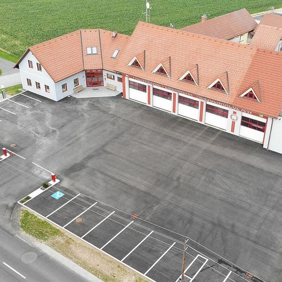 Eine Draufsicht auf eine Feuerwache mit rotem Dach und einem großen Parkplatz davor. Vor dem Eingang stehen Bänke.