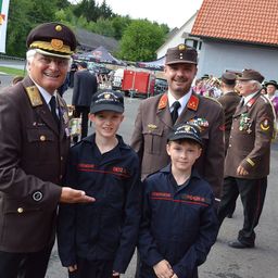 Drei Männer in Militäruniformen stehen neben zwei Jungen in passenden Uniformen. Die Männer tragen Mützen mit Abzeichen und die Jungen haben Mützen mit der Aufschrift Feuerwehr. Sie stehen vor einem Gebäude mit rotem Dach und einer Gruppe von Menschen im Hintergrund.