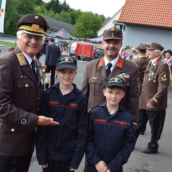 Drei Männer in Militäruniformen stehen neben zwei Jungen in passenden Uniformen. Die Männer tragen Mützen mit Abzeichen und die Jungen haben Mützen mit der Aufschrift Feuerwehr. Sie stehen vor einem Gebäude mit rotem Dach und einer Gruppe von Menschen im Hintergrund.