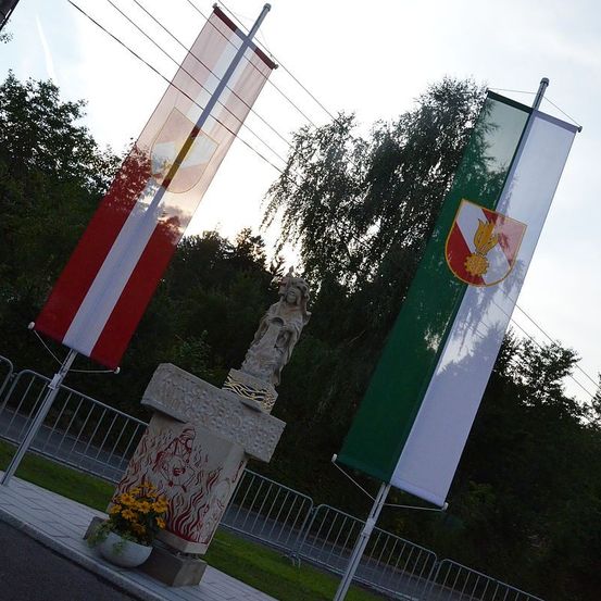 Ein Denkmal steht in einem Park mit zwei Flaggen. Eine Flagge ist rot und weiß, die andere grün und weiß. Es gibt eine Statue und eine gelbe Blume in einem Topf. Bäume und ein Zaun sind im Hintergrund.