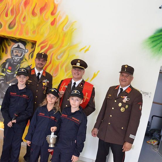 Eine Gruppe von Feuerwehrleuten und Kindern in Uniformen steht vor einem Feuerwehrmann-Wandbild. Die Kinder halten einen silbernen Pokal.
