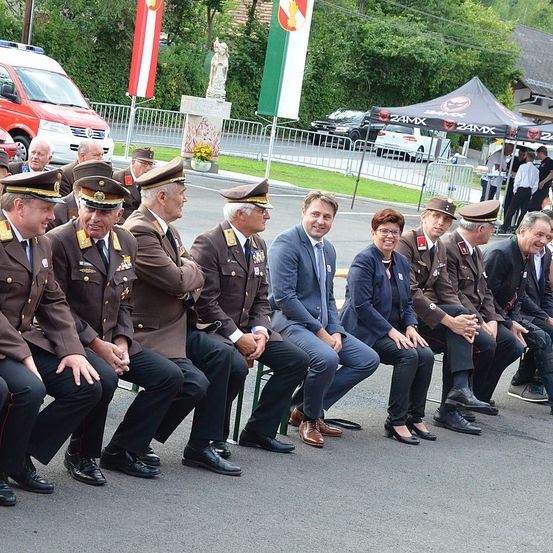 Eine Gruppe von Militärangehörigen und ein Paar in Zivilkleidung sitzen im Freien. Hinter ihnen befinden sich Flaggen, eine Statue und ein Zelt. Autos und ein Krankenwagen sind am Straßenrand geparkt.