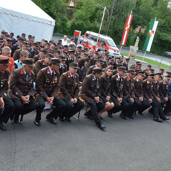 Eine Gruppe von Männern in Militäruniformen sitzt auf dem Boden, wahrscheinlich bei einer Parade. Sie tragen alle Mützen, Schuhe und Medaillen. Hinter ihnen steht ein weißes Zelt, und in der Ferne sind einige Autos und Flaggen zu sehen.