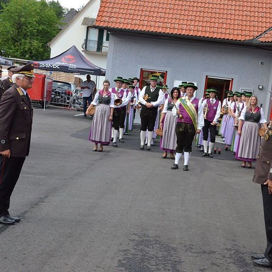 Eine Gruppe von Menschen in traditionellen Kostümen, möglicherweise für einen Umzug, geht auf der Straße. Ihnen folgen zwei Offiziere. Dahinter ist ein Gebäude mit rotem Dach und einem Zelt zu sehen.