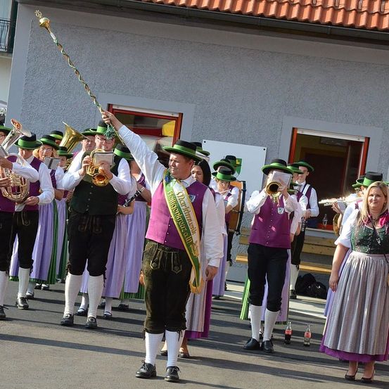Eine Blaskapelle in traditioneller deutscher Kleidung tritt im Freien auf, wobei die Musiker verschiedene Instrumente spielen, darunter Trompete und Posaune.