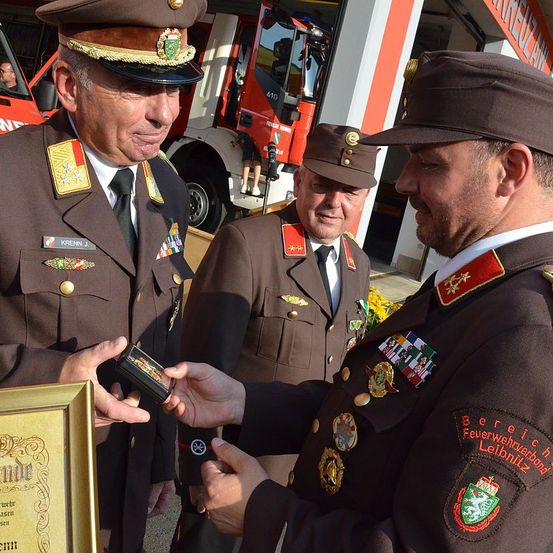 Drei Männer in Militäruniformen stehen und tauschen Gegenstände aus. Der Mann auf der linken Seite hält ein gerahmtes Zertifikat, während der Mann auf der rechten Seite ein kleines Objekt in der Hand hat. Sie stehen in der Nähe eines Feuerwehrwagens und eines Gebäudes.