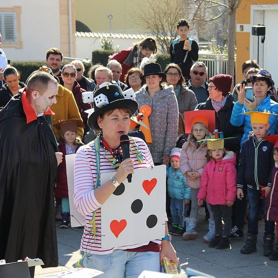 Eine Frau mit Mikrofon und einem Schild mit Würfelgesicht spricht zu einer Menge von Erwachsenen und Kindern, einige in Hüten und Kostümen gekleidet.