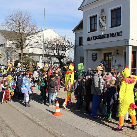 Eine Gruppe von Menschen in Kostümen steht vor einem Gebäude. Einige tragen Hüte und Schuhe. Eine Person in einem Hühnerkostüm steht neben einem Verkehrskegel. Bäume und Gebäude im Hintergrund.